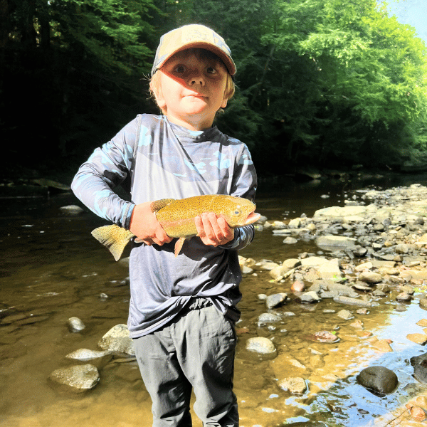 Young boy in grey HELIOS sun shirt holding trout by river wearing cap and sunglasses