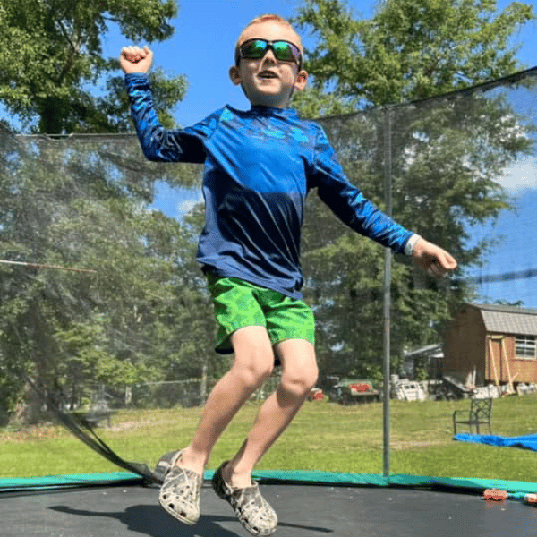 Young boy jumping on trampoline wearing blue and black HELIOS sun shirt and green shorts