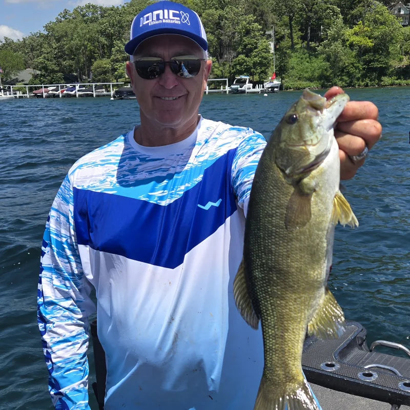 Angler wearing Helios long-sleeve performance fishing shirt on boat with bass catch in bright sunny conditions, demonstrating superior UPF 50+ sun protection and professional fishing performance
