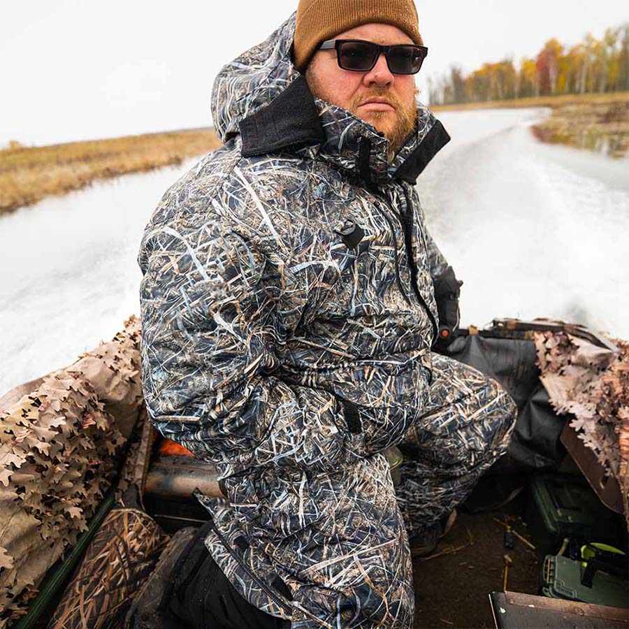 Waterfowl hunter in a WindRider Hayward Wetlands Camo float jacket riding a running duck boat through a marsh channel on an overcast late-season morning
