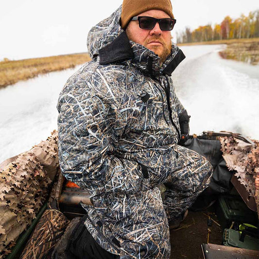 Waterfowl hunter in a WindRider Hayward Wetlands Camo float jacket riding a running duck boat through a marsh channel on an overcast late-season morning