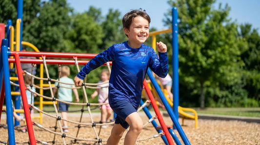 kid around age 7 running across a sunny playground climbing structure at midday, wearing a long-sleeve UPF sun shirt, other kids blurred in background, bright natural light