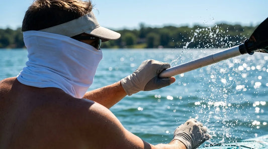 kayaker on a sunlit lake mid-paddle-stroke, water droplets flying off the paddle blade, UPF sun glove visible gripping the shaft, bright midday light reflecting off the water surface
