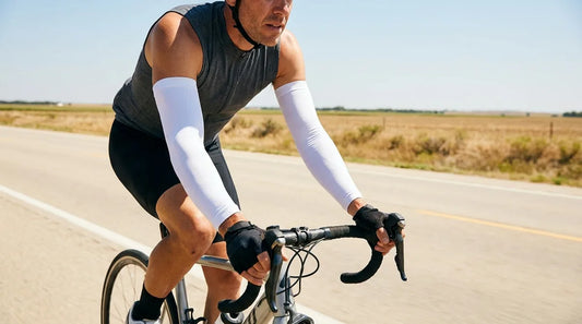 road cyclist on a sunlit two-lane highway shoulder, wearing fitted white UPF arm sleeves over bare shoulders, hands on drop bars, midday sun casting hard shadows