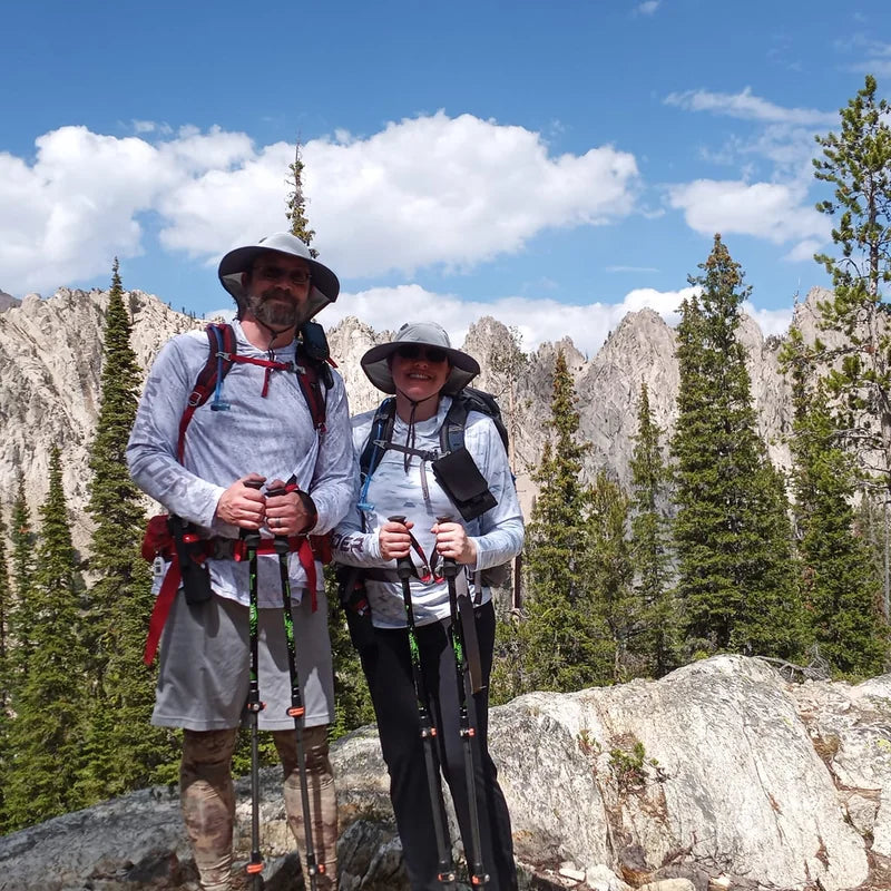Couple at alpine summit wearing WindRider Helios sun protection shirts with backpacks and rocky mountain peaks in background
