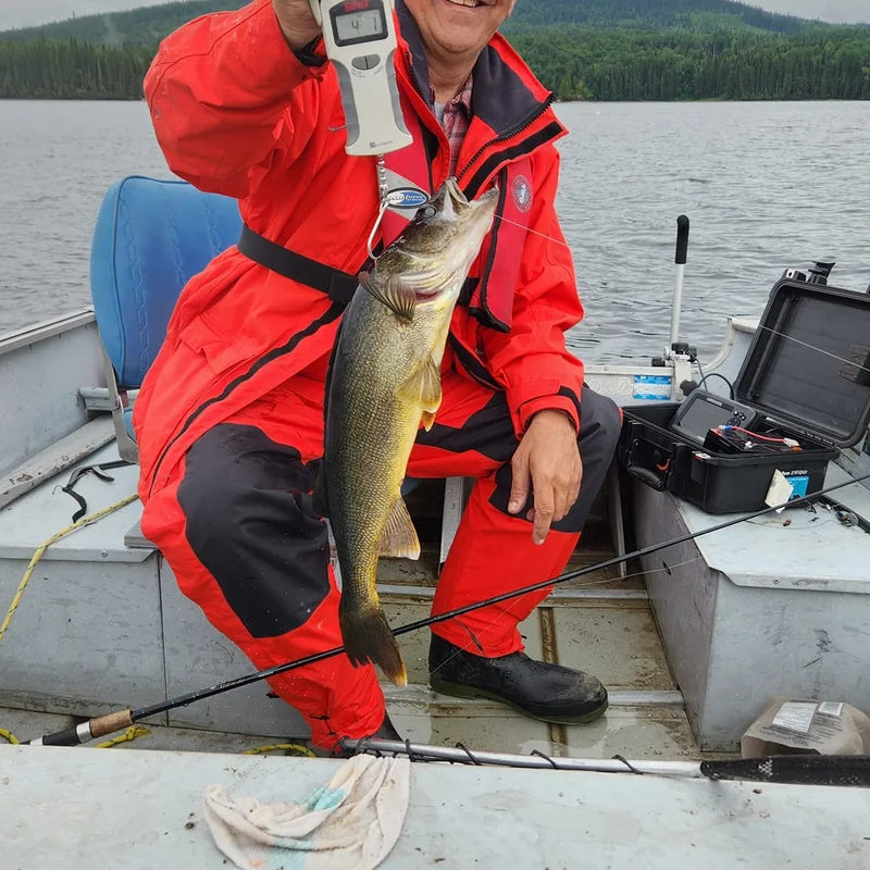 Angler wearing red rain suit jacket and bibs sitting in aluminum boat on overcast lake holding a walleye