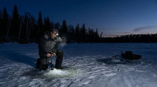 angler kneeling on dark early-season ice at dusk, drilling a hole with an auger, headlamp on, dark water showing through clear thin ice nearby, winter treeline in background