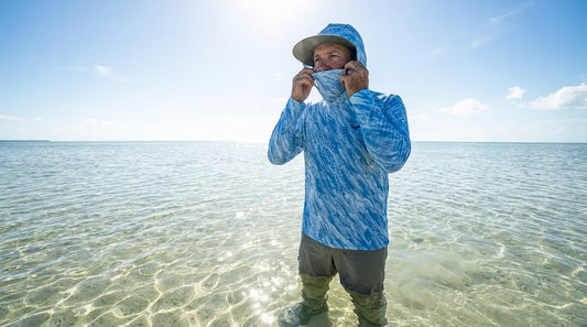 angler in open flats water pulling up integrated gaiter on hooded sun shirt, bright overhead sun, clear shallow water visible