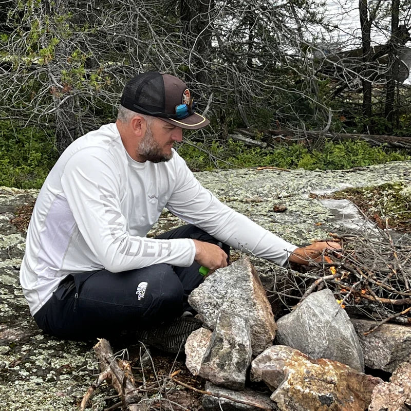 Man in white Helios sun shirt working outdoors building campfire on rocky shore