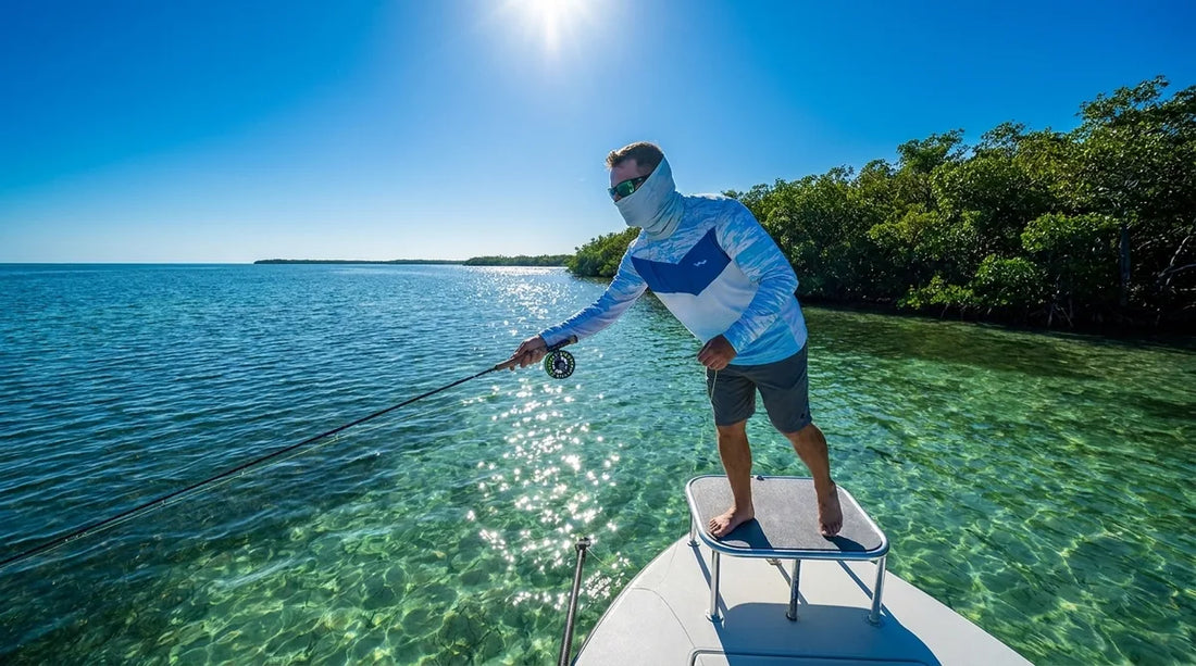 angler on open flats boat in bright Florida summer sun, fishing for redfish or snook, wearing light-colored long-sleeve UPF shirt and sun gaiter pulled up, blue sky and mangrove shoreline in background