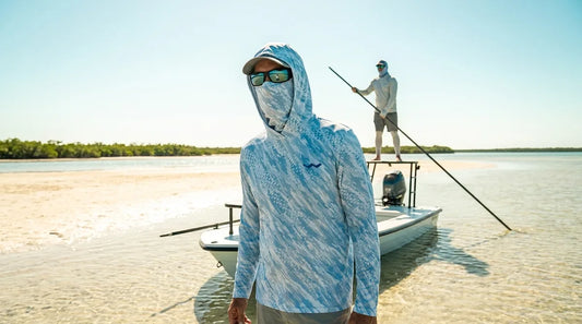 angler standing on the bow platform of a poling skiff on skinny water, hooded sun shirt with gaiter pulled up over nose and mouth, poling pole angler visible behind on platform, harsh midday light reflecting off pale sand flats