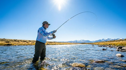 fly fisherman wading knee-deep in a clear mountain stream, casting with full arm extension in bright midday sun, wearing long-sleeve UPF shirt and fingerless sun gloves, surrounded by open treeless riverbank