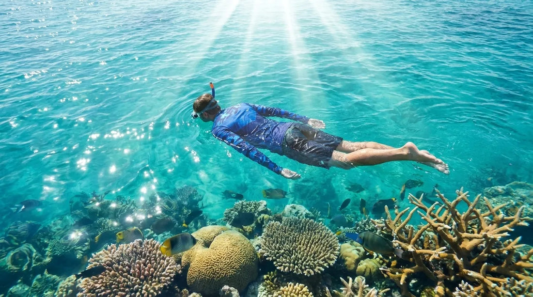snorkeler floating face-down at the surface in clear tropical water, wearing a long-sleeve UPF sun shirt over boardshorts, bright midday sun, coral reef visible below