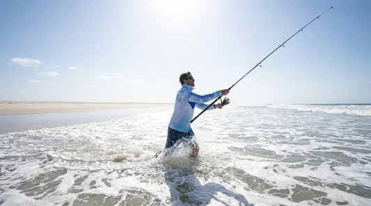angler in long-sleeve UPF shirt wading in waist-deep surf, casting rod with bright midday sun and white-capped waves, sand and open beach visible behind