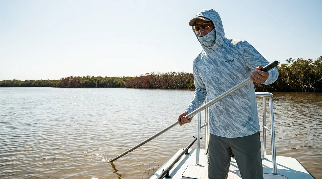 angler standing on the raised poling platform of a flats skiff scanning a backcountry mangrove flat for tarpon, harsh midday sun overhead, fully covered in a hooded sun shirt with gaiter pulled up over the nose
