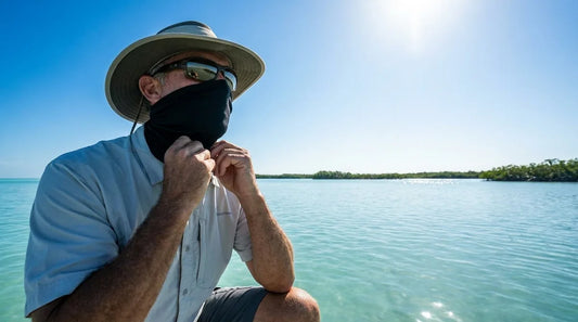 angler on a saltwater flat adjusting a neck gaiter pulled up over nose and chin, bright midday sun, calm blue water in background