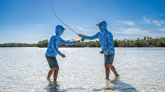 angler on bay flat in bright summer sun, wearing a hooded UPF shirt with integrated gaiter pulled up over nose and chin, rod in hand mid-cast, water reflecting sunlight