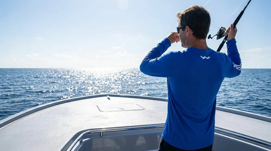 angler on a bright, sun-drenched boat deck at midday casting a line, wearing a long-sleeve blue UPF sun shirt, water glare visible in the background