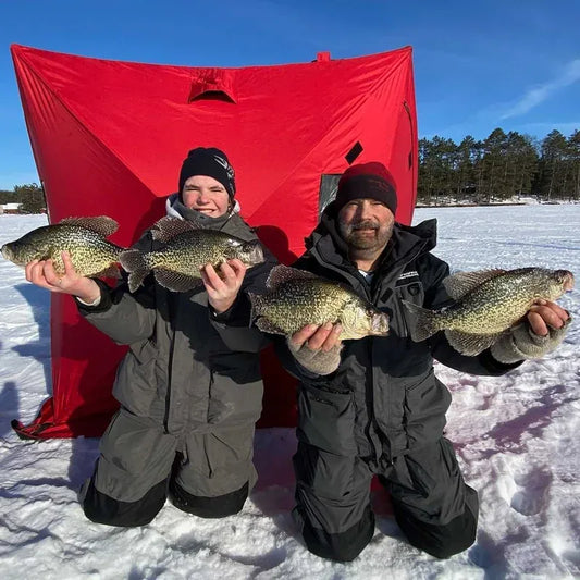 Two anglers kneeling in grey WindRider Boreas ice fishing bibs beside a red pop-up shelter on a sunny frozen lake, holding four large crappie
