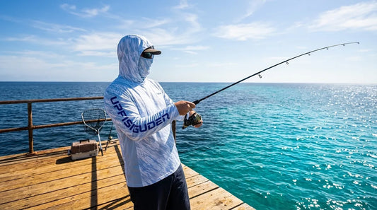 angler standing at a wooden ocean pier railing, midday sun high overhead, casting a rod with open water stretching to the horizon, wearing a long sleeve hooded UPF fishing shirt with gaiter pulled up, light reflecting off the water surface below
