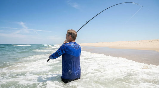 surf angler standing in waist-deep ocean water casting a long rod into breaking waves, bright summer sun directly overhead, open beach stretching behind them with no shade in sight