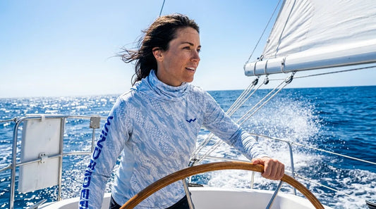 woman at the helm of a sailboat, hand on the wheel, spray coming over the bow, bright midday sun and glare off blue water