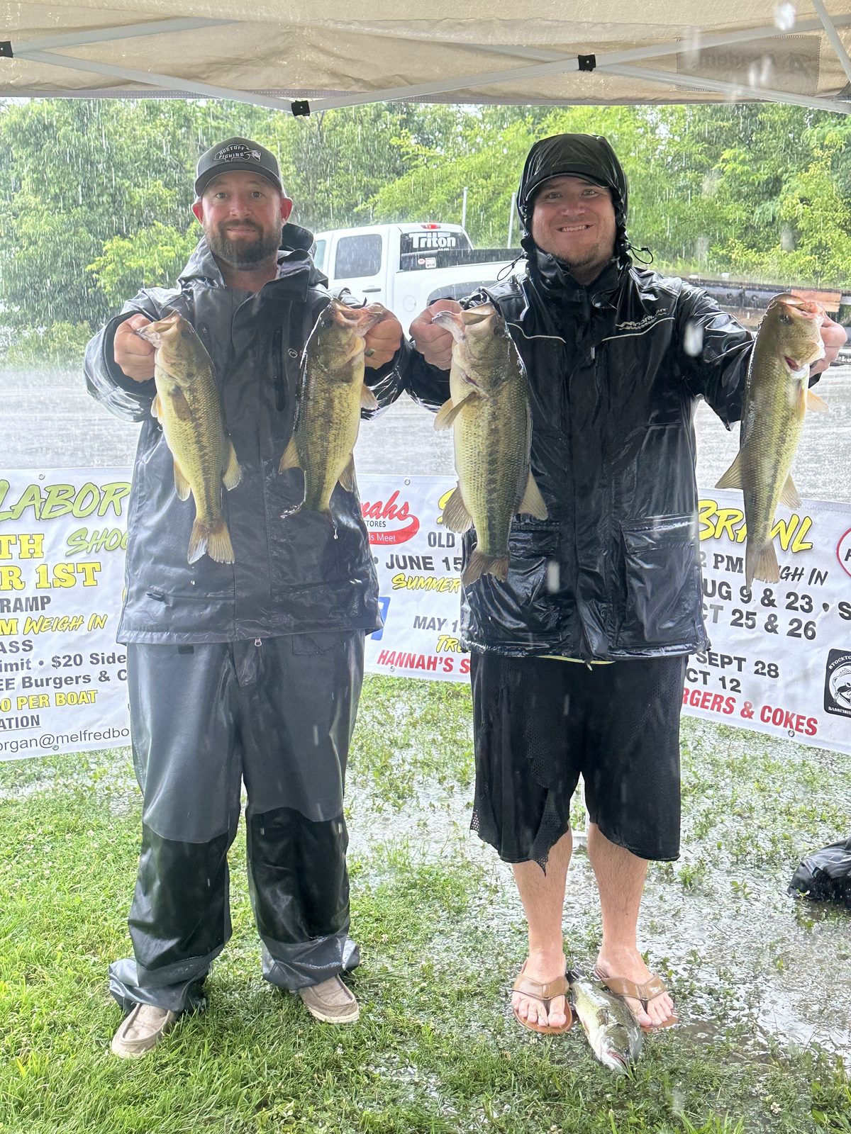 Two fishermen at a rainy tournament weigh-in holding bass, both wearing black Pro All-Weather jackets and bibs