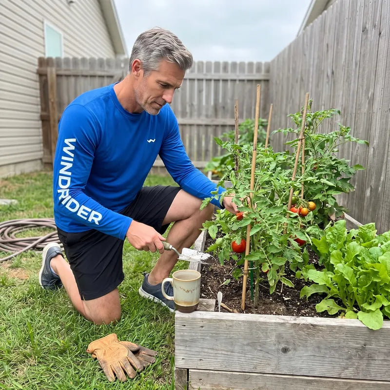A man kneeling in his backyard vegetable garden wearing the blue Helios sun shirt.
